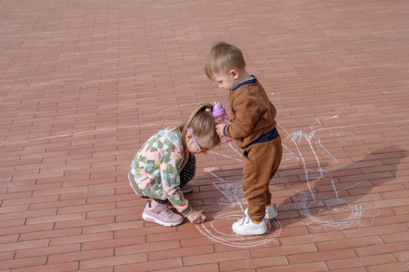 Girl with her younger brother doodling with chalk on pavement, overhead view