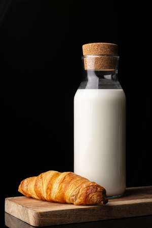 Bottle with milk on a wooden board. Black background. Glass bottle. Natural cork. White drink. Cows milk. One classic french croissant on a white background. Homemade baking.の写真素材