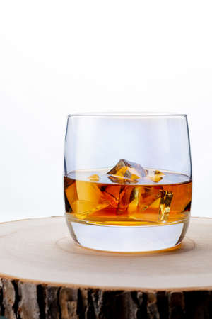 Glass of whiskey with ice on a wooden board. Wooden saw cut. White background. Whiskey / Brandy / Cognac / Rum. Glass with a strong drink.の写真素材