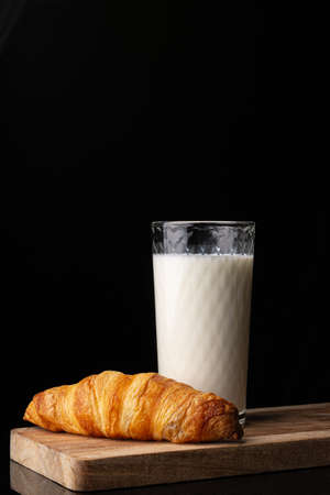 A glass with milk and one french croissant on a wooden board. Black background. White drink. Cows milk. Classic french croissant. Homemade baking.の写真素材