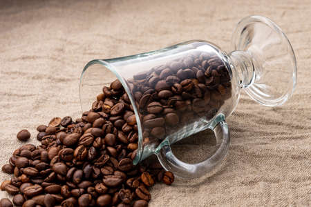 Coffee beans sprinkled from a transparent glass goblet. Fried arabica. Glass for latte. Glass for mulled wine. Linen background. Sackcloth. Nettle tissue.の写真素材