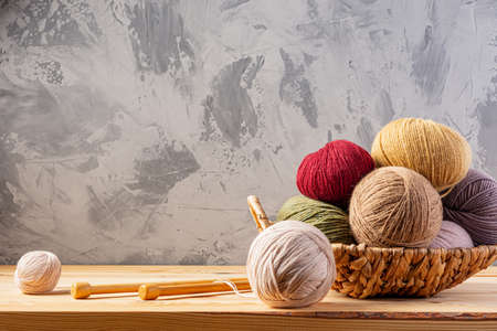 Skeins of yarn for knitting from natural wool in a wicker basket. Wooden surface or table. Background - concrete wall. Wooden knitting needles.の写真素材