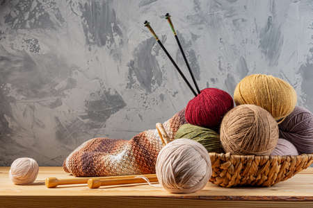 Skeins of yarn for knitting from natural wool in a wicker basket. Wooden surface or table. Background - concrete wall. Wooden knitting needles.の写真素材