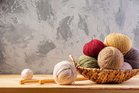 Skeins of yarn for knitting from natural wool in a wicker basket. Wooden surface or table. Background - concrete wall. Wooden knitting needles.の写真素材