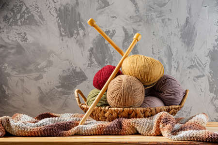 Skeins of yarn for knitting from natural wool in a wicker basket. Wooden surface or table. Background - concrete wall. Wooden knitting needles.の写真素材