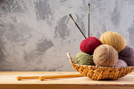 Skeins of yarn for knitting from natural wool in a wicker basket. Wooden surface or table. Background - concrete wall. Wooden knitting needles.の写真素材