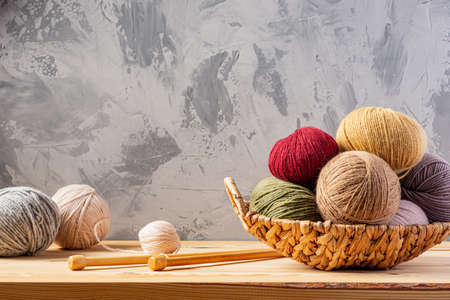 Skeins of yarn for knitting from natural wool in a wicker basket. Wooden surface or table. Background - concrete wall. Wooden knitting needles.の写真素材
