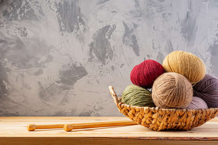 Skeins of yarn for knitting from natural wool in a wicker basket. Wooden surface or table. Background - concrete wall. Wooden knitting needles.の写真素材