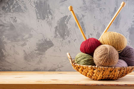 Skeins of yarn for knitting from natural wool in a wicker basket. Wooden surface or table. Background - concrete wall. Wooden knitting needles.の写真素材