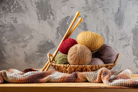 Skeins of yarn for knitting from natural wool in a wicker basket. Wooden surface or table. Background - concrete wall. Wooden knitting needles.の写真素材