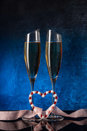 Two glasses with champagne on a black mirror surface. Heart for St. Valentine. Colored satin ribbon. Blue background Concrete wall. Black glass. Crystal glassの写真素材