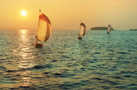 Three sailing yachts on the background of the setting sun, visible in the distance the shore and the...