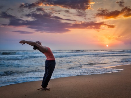 Young sporty fit woman doing yoga Sun salutation Surya Namaskar pose Hasta Uttanasana on tropical beach on sunsetの写真素材