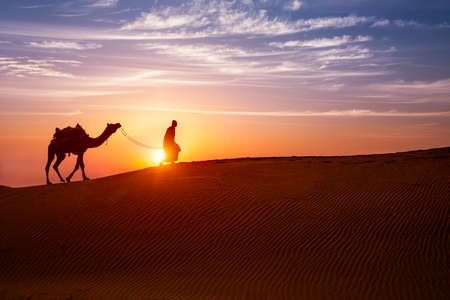 Indian cameleer camel driver with camel silhouettes in dunes on sunset. Jaisalmer, Rajasthan, Indiaの写真素材