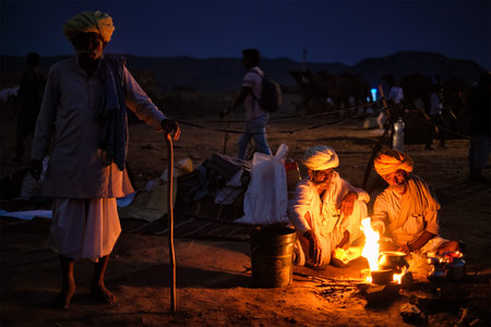 Pushkar, India - November 6, 2019: Indian rural village men and camels lit by camp fire in night at...