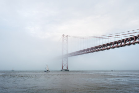 View of 25 de Abril Bridge famous tourist landmark of Lisbon connecting Lisboa and Almada in heavy fog mist wtih yacht boats passing under. Lisbon, Portugalの写真素材