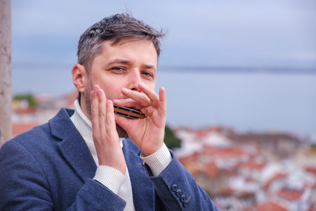 Man musician in a blue blazer and white sweater playing a harmonica with eyes closed, standing outdoors in city streetの写真素材