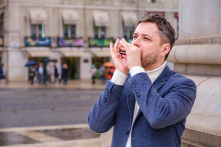 Man musician in a blue blazer and white sweater playing a harmonica with eyes closed, standing outdoors in city streetの写真素材