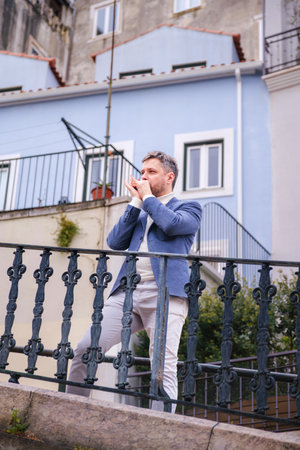 Man musician in a blue blazer and white sweater playing a harmonica with eyes closed, standing outdoors in city streetの写真素材
