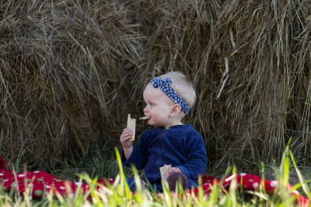 Little baby girl is sitting by the haystack and eating a crisp bread.の写真素材