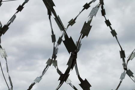 Closeup shot of a barbed steel wire fence with cloudy sky on the background.