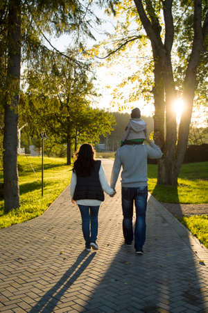 Family is walking in park at sunset, mother and father are holding hands, son is sitting on shoulders of his father.の写真素材