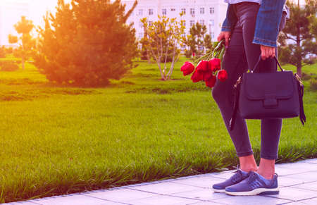 Young girl in jeans with bouquet of tulips flowers and bag walks in park with green grass. Love and lifestyleの写真素材