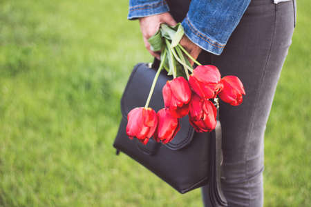 Young girl in jeans with bouquet of tulips flowers and bag walks in park with green grass. Love and lifestyleの写真素材