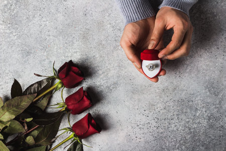 Valentines day romantic man hand holding engagement ring in box marry me wedding with red roses bouquet gift surprise on grey background. Love gift for woman making proposalの写真素材
