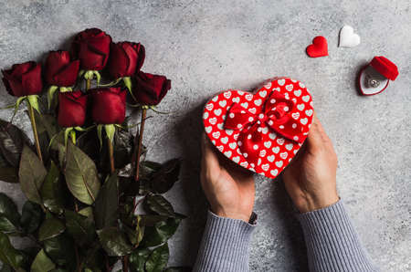 Valentines day man hand holding gift box heart shape mothers day red rose marry me wedding engagement ring in box surprise on grey background. Love flower gift for woman on romantic holiday birthdayの写真素材