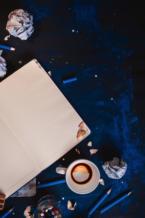Open notepad with blank pages, crumpled paper balls, pencils, notepads and empty coffee cups on a dark background. Still life with writer workplace. Creative writing concept.の写真素材