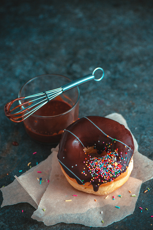Chocolate donut with sprinkles on dark background with a whisk and chocolate glazing. Low key food photography. Wax baking paper.の写真素材