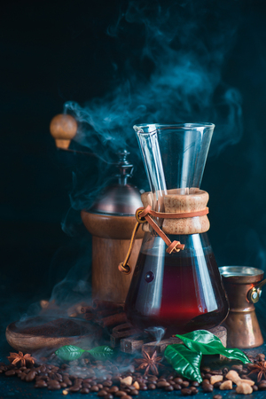 Hourglass coffee maker with a coffee grinder, little cezve, beans, anise stars, brown sugar, and Arabica leaves on a dark background with steam. Alternative coffee brewing method. Drink photography with copy space.の写真素材