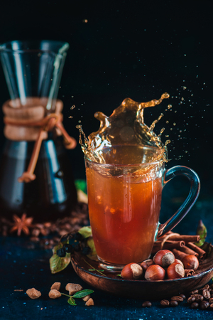 Coffee splash in a tall latte glass with hourglass coffee maker, spices, nuts and coffee beans on a dark background. Action drink photography.の写真素材