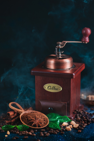 Coffee grinder with roasted coffee beans and steam on a dark background. Arabica leaves, brown sugar and anise stars as spices. Vintage still life with hot drink.の写真素材
