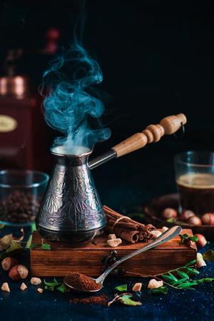 Steaming cezve with hot coffee on dark background with beans, cinnamon, nuts, leaves and coffee grinder. Brewing coffee concept.の写真素材