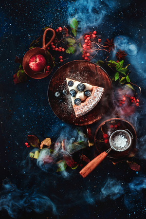 A piece of cake with powdered sugar and a strainer on a dark wooden background with floral decoration. Handmade wooden plates in autumn flat lay. Conceptual stylised food still life, food and drink table top shot on dark background.の写真素材