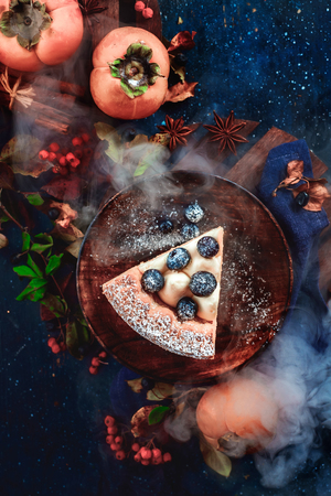 Close-up of a piece of pumpkin pie with blueberries and persimmon on a dark wooden background with floral decoration. Handmade wooden plates in autumn flat lay. Conceptual stylised food still life, food and drink table top shot on dark background.の写真素材