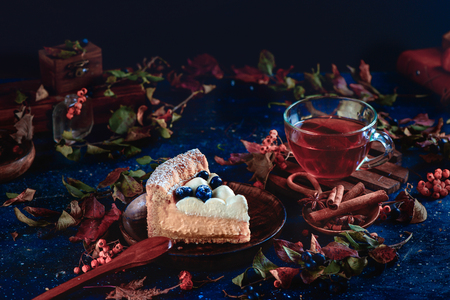 Hommemade crust cake with wipped cream, blueberries and pumpkin puree on a dark background with teacup, autumn leaves and cinnamon. Close-up of a piece of cake. Dark food photography. Autumn still life with pastry.の写真素材