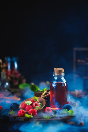 Raspberry potion with a wooden bowl of berries and mint leaves on a dark background. Magical still life with a mystic brew. Summer vibes and preserves for winter. Smoke and copy space.の写真素材