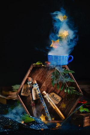 Test tubes with potion ingredients, lavender, berries, and stardust. Magical still life with staming cup of herbal tea, hexagonal wooden tray and dark background. Fairy tale concept with copy space.の写真素材