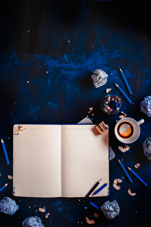 Open notepad with blank pages, crumpled paper balls, pencils, notepads and empty coffee cups on a dark background. Still life with writer workplace. Creative writing concept.の写真素材