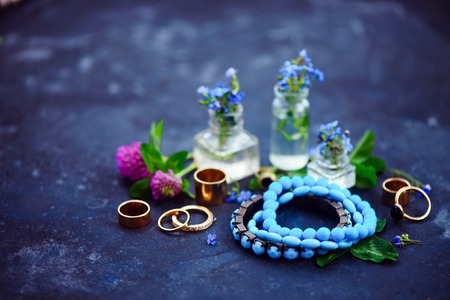 Blue bead bracelets and golden rings in a feminine style concept with blue flowers in tiny bottles on a dark background. Beautiful summer accessories with copy spaceの写真素材