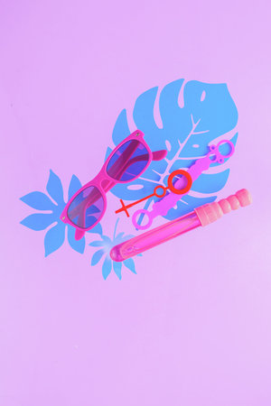 Bubble wands and sunglasses on a pastel purple background with tropical leaves and copy space....