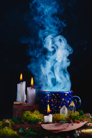 Magical ceramic cup with constellations and stars in a dark still life. Witch or astrologer workplace with burning candles, smoke, crystals, moss and potion bottles. Magical concept with copy spaceの写真素材