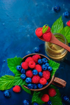 Raspberry and blueberry in a ceramic cup with green leaves and a glass jar of honey. Summer berry harvest concept on a dark blue wet background with copy space. Top view food photographyの写真素材