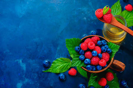 Raspberry and blueberry in a ceramic cup with green leaves and a glass jar of honey. Summer berry harvest concept on a dark blue wet background with copy space. Top view food photographyの写真素材