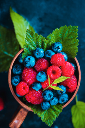 Raspberry and blueberry with green leaves in a ceramic cup. Summer berry harvest concept on a dark blue wet background with copy space. Top view food photographyの写真素材