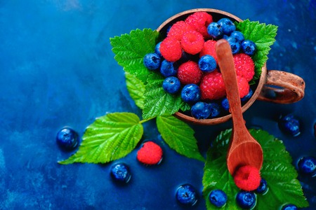 Colorful assorted mix of raspberry and blueberry with green leaves in a ceramic cup. Summer vitamins concept on a dark blue wet background with copy spaceの写真素材