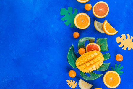 Exotic fruits flat lay. Mango, oranges, kumquat and other tropical fruits on a monstera plate. Bright blue background with copy space.の写真素材